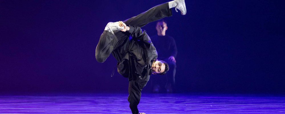 ABC (Academy Breakin’ Convention) A dancer dressed in all black balances in a one-handed handstand