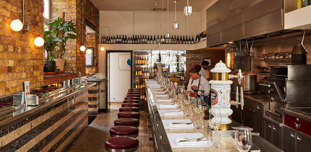 A modern restaurant kitchen featuring an open counter with red stools, place settings, and a chef preparing food. Wine bottles are displayed on shelves, and plants adorn brick and tiled walls.