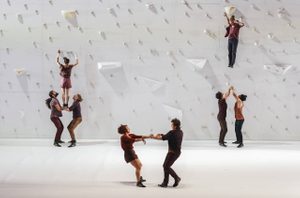 Corps extrêmes © Pascale Cholette A giant white rock climbing wall. In the background are two groups of three. Two bases and one flyer mid-air. In the foreground, two dancers are on their feet, holding hands as they move