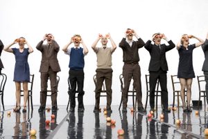 Gandini Juggling - Smashed © Ludovic des Cognets A group of jugglers in a big white room stand in front of chairs and hold up apples to their eyes