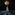 A black man with a shaved head wearing a print cotton dress steps away from us. A huge image of a moon, almost as tall as him, floats overhead. The black stage is raked with silvery lines of moonlight, and dim figures lie on the floor in the distance.
