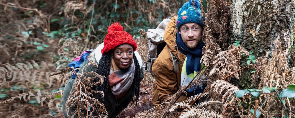 A man and a woman in woolly hats are crouched behind foliage on the forest floor, peering out excitedly at something they have seen ahead.