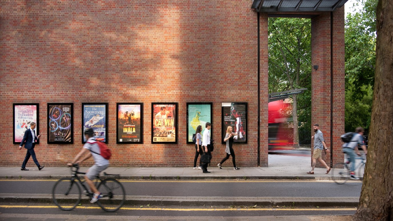 A photo of the outside wall of Sadler's Wells Theatre building with show posters and pedestrians and a cyclist passing by