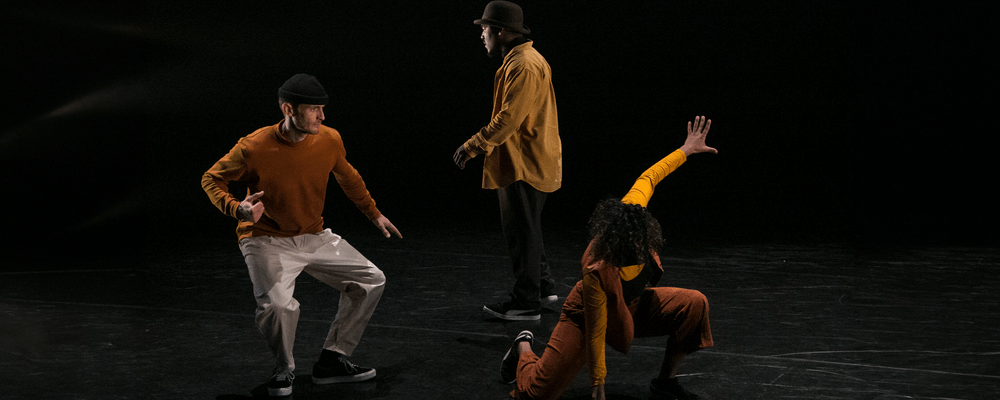Back to the Lab 2025 © Dave Barros Three people perform a dynamic dance on a dark stage; one kneels with an arm raised, another stands facing forward, and the third balances on one leg.