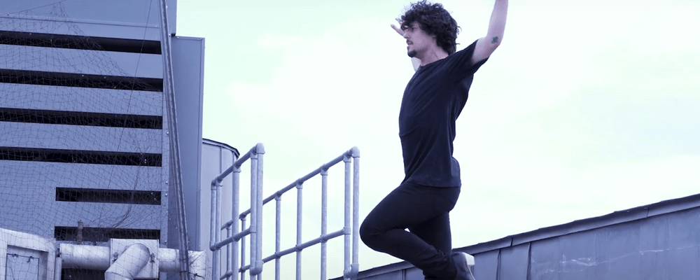 Jesús Carmona A photograph of Jesús Carmona performing a flamenco move, with his left up arm and his right arm to the side. He has dark curly hair and is wearing a black t-shirt.
